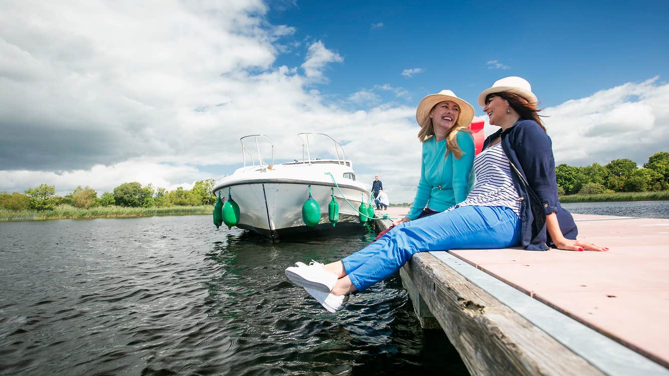 Two people wearing hats relaxing beside a river and boat in Portumna, Co. Galway in Ireland's Hidden Heartlands.