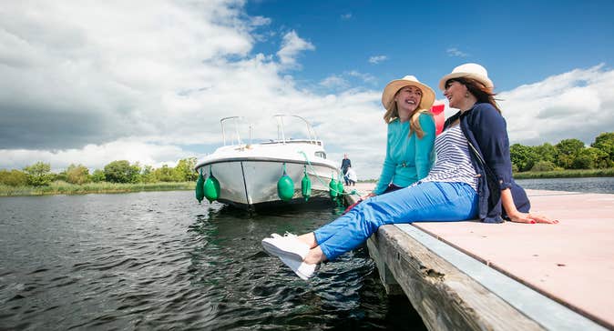 Two people wearing hats relaxing beside a river and boat in  Portumna, Co. Galway in Ireland's Hidden Heartlands.