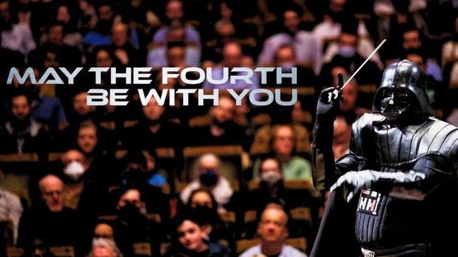 A figure in black shiny darth vadar outfit with a conductor's baton against background of an audience