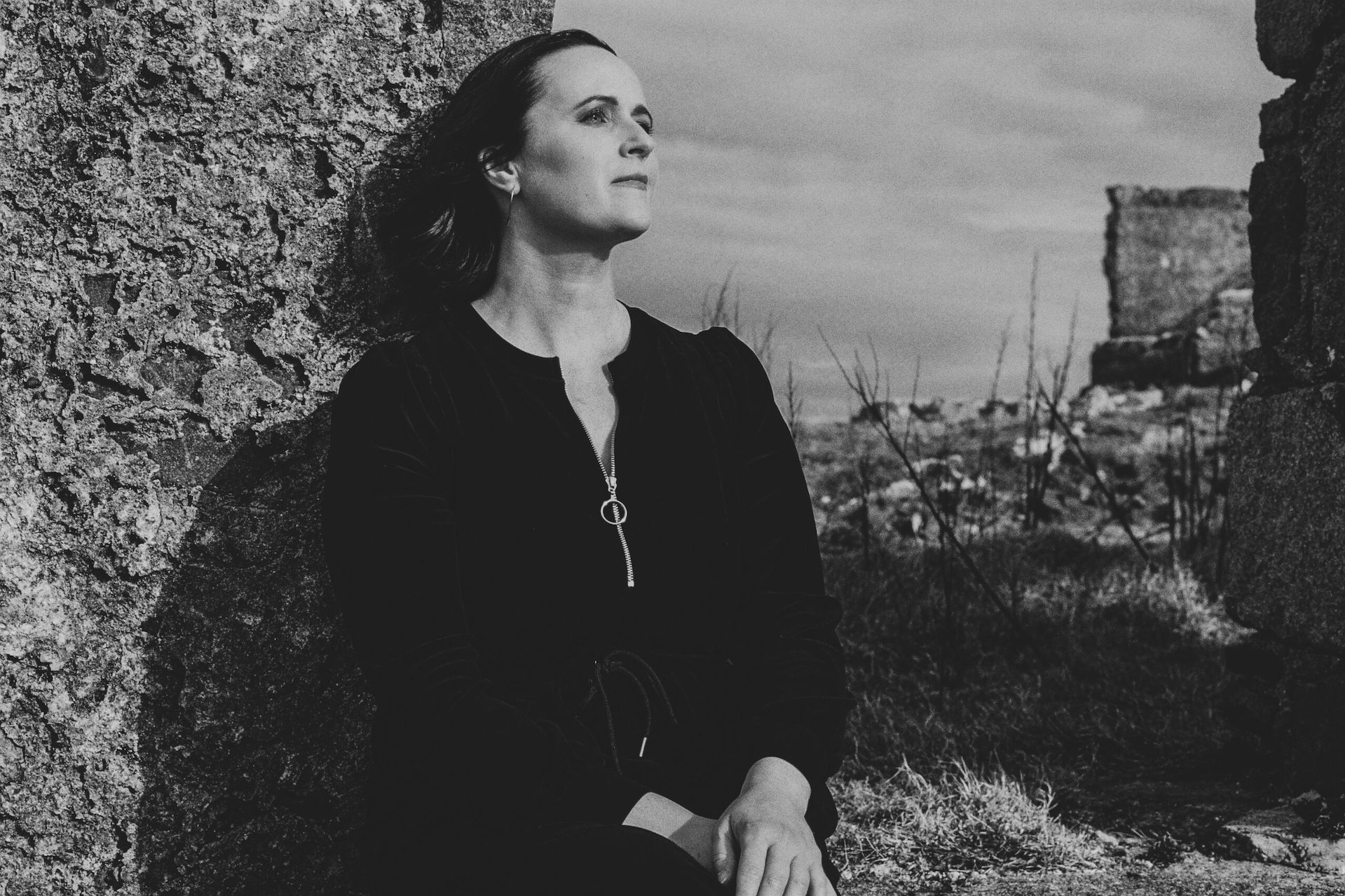 Black and white photo of woman standing outside looking away into the distance with partial stone wall and barren landscape behind her.