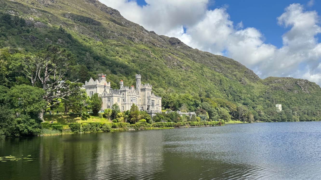 Abbey nestled among trees and mountains with a lake in front of it