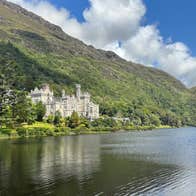 Abbey nestled among trees and mountains with a lake in front of it