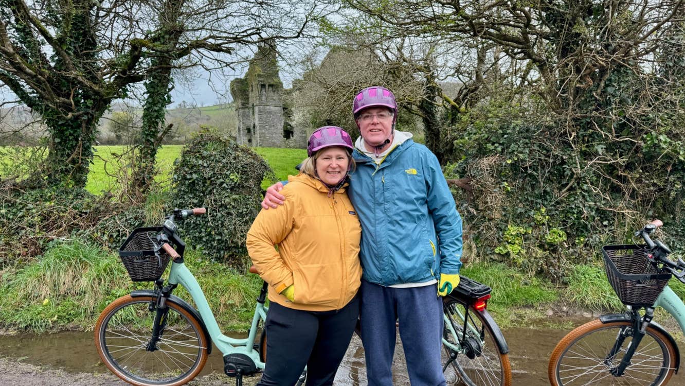 A couple pose for a photo with their eBikes on a country lane near an old ruin