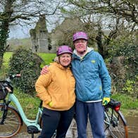 A couple pose for a photo with their eBikes on a country lane near an old ruin
