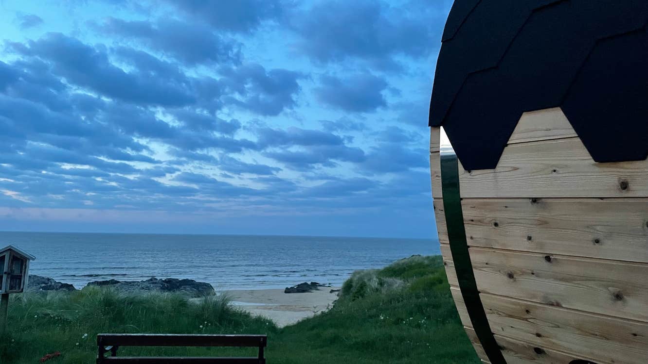 Wooden sauna next to a bench overlooking a beach and grass in the evening time