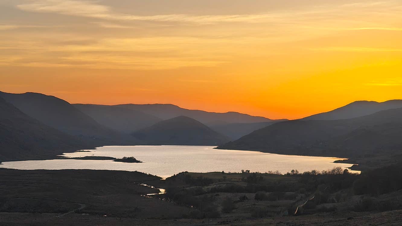 Sunset over Lough Nafooey with golden light reflecting on the lake, surrounded by layered mountain silhouettes and rolling hills in Joyce Country.