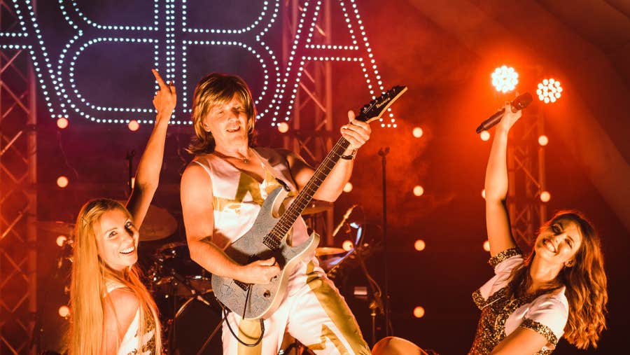 On a red lit stage a man is playing an electric guitar with women smiling and kneeling either side.