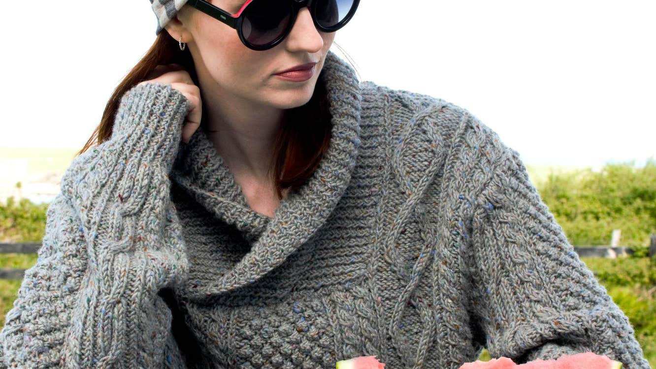 Person wearing sunglasses a headscarf and grey woollen jumper sitting at a bench with a sliced watermelon in front