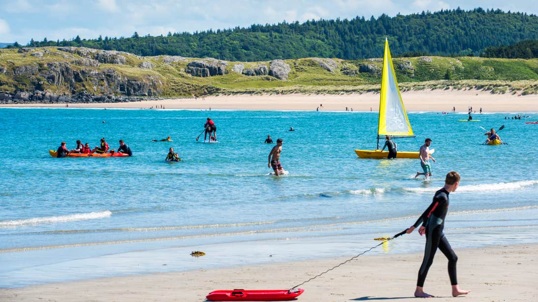 Man pulling a red board along the sand in front of people playing in the sea at Marble Hill Beach, Donegal