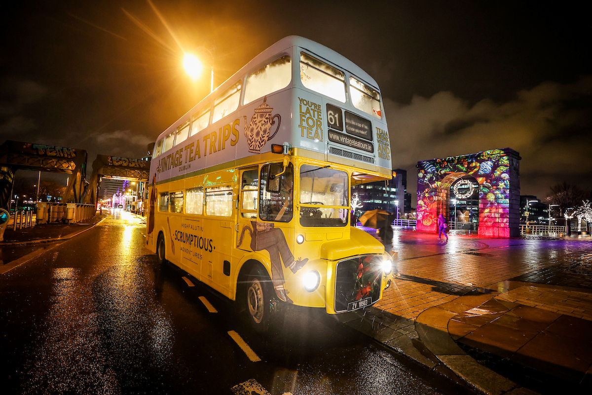 An old fashioned, double decker bus in yellow and light blue, parked up at night in a city street.