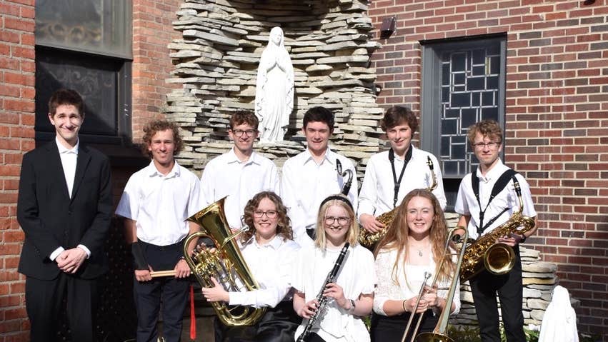 A small group of musicians, some holding instruments, in front of a statue of Our Lady.
