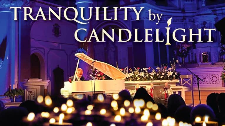 From a distance, a man seated at a large, pale coloured grand piano with flowers in the background and blurry candles in the foreground.