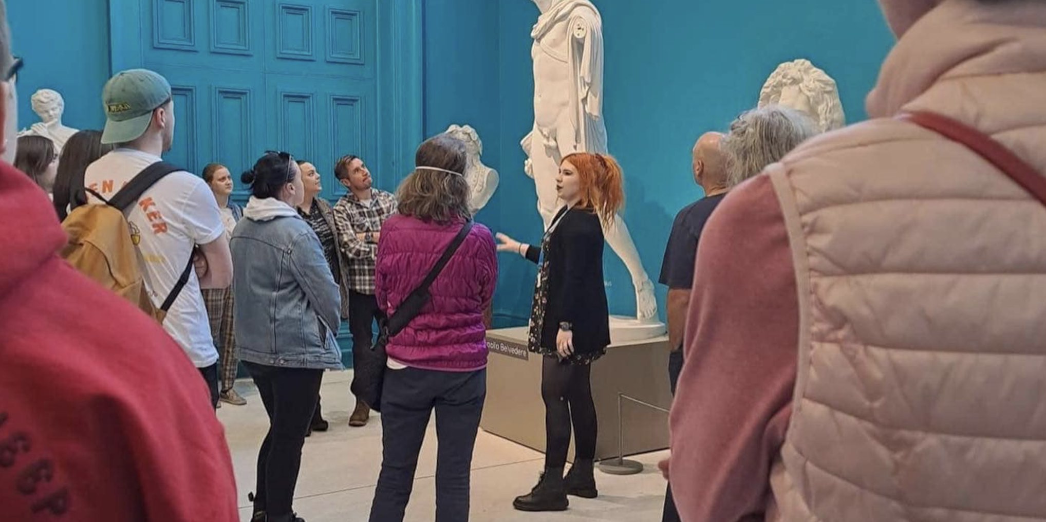 A tour guide is standing in front of a tall, white, stone statue in a blue room talking to a group of people.
