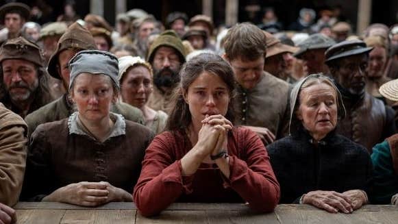 A worried woman resting her elbows on wooden stage at the front of crowd of sad looking people in period costumes.