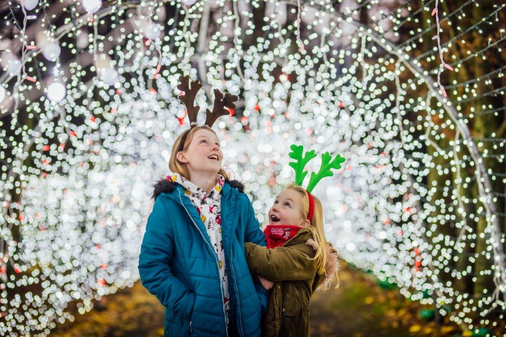 2 young girls with reindeer antlers on heads are looking with delight at the tunnel of thousands of white lights they are standing in.