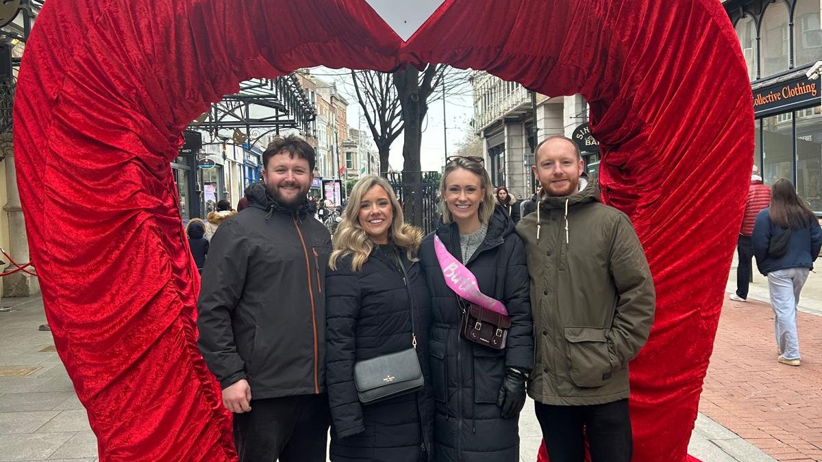 Friends pose beneath a giant red velvet heart arch in the heart of Dublin, part of the city's playful embrace of romance and love around Valentine's season, with the Gaiety Theatre visible in the background.