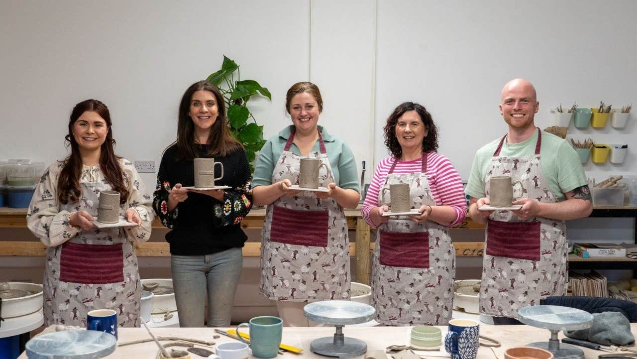 Five people with their pottery pieces standing at a table with aprons on them