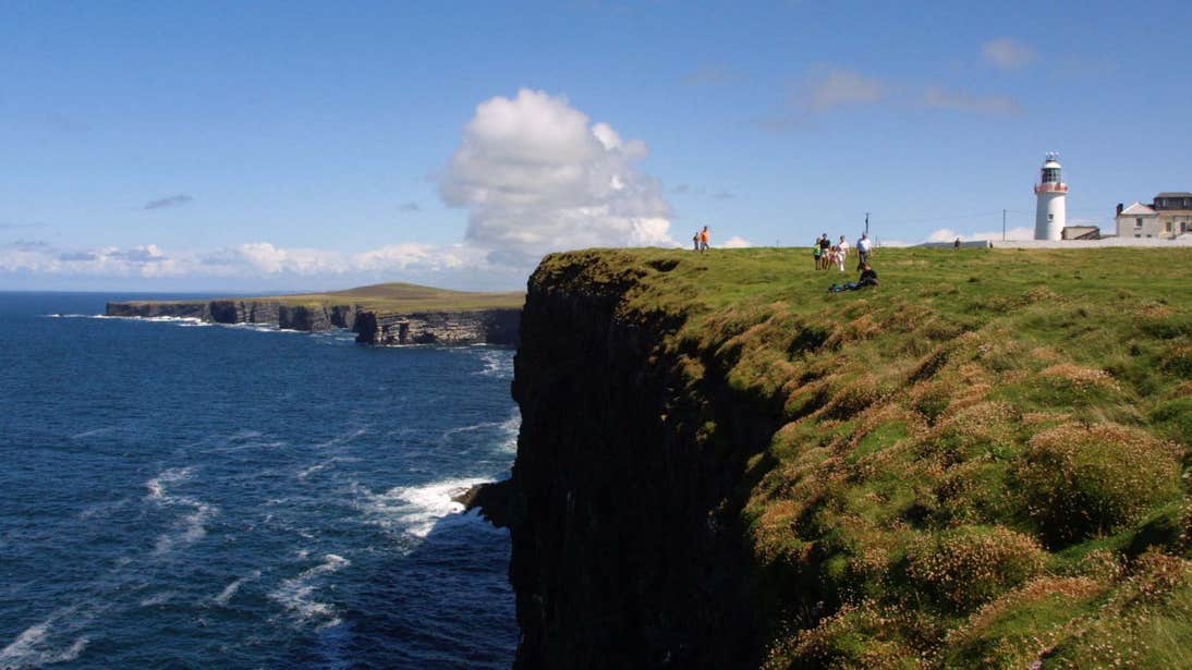People walking on a trail beside a cliff with a lighthouse in the distance at Loop Head, Clare