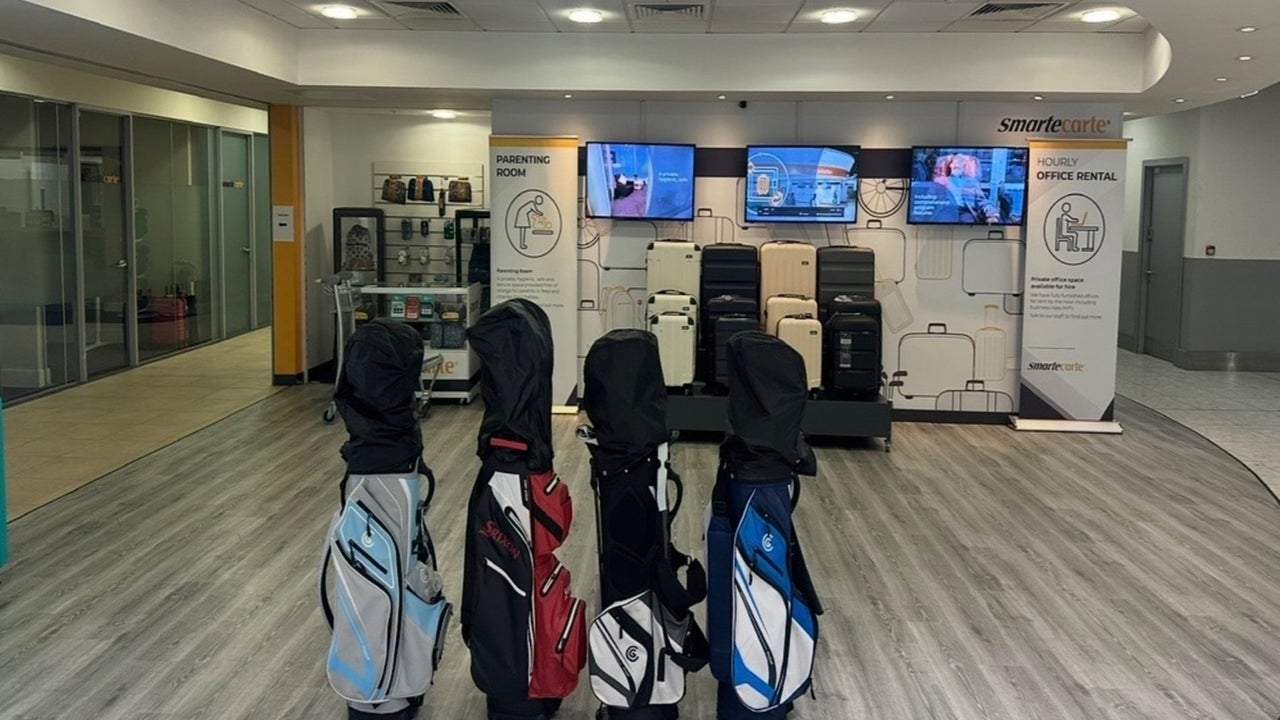 Four golf club bags standing on a light wooden floor with three screens and suitcases in the background