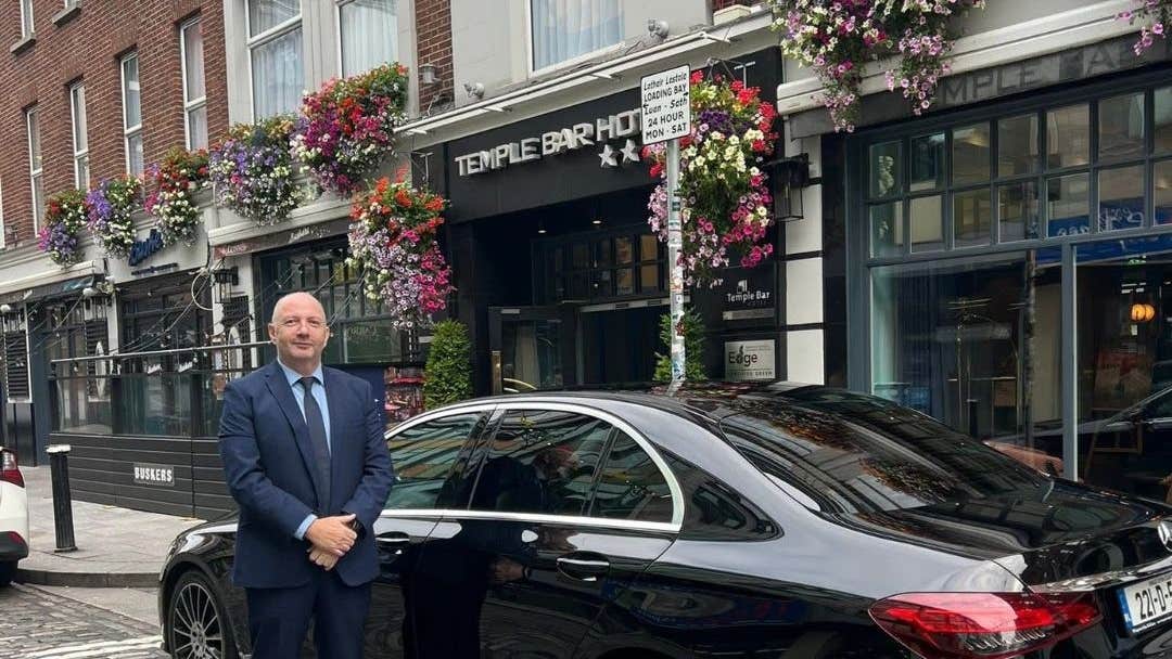 Person in a suit standing next to a black car parked on a street outside a hotel with flowers