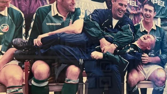 A group of laughing men in football strips seated with another man laid horizontally across their laps also laughing.