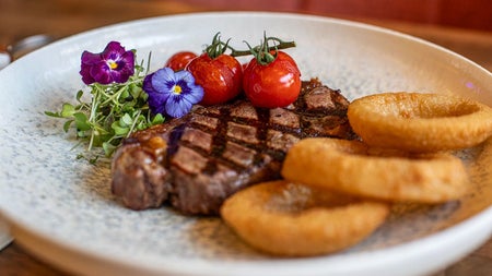 A cooked steak on a white plate with vine tomatoes, flowers and onion rings