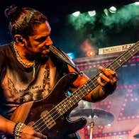 A man in leather trousers and gold jewelry is playing an electric guitar on a stage