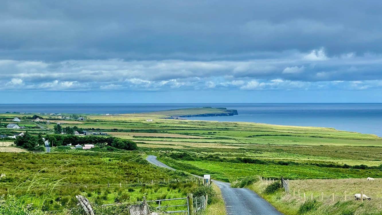 A country road meanders through green countryside with a cloudy sky and the sea in the distance