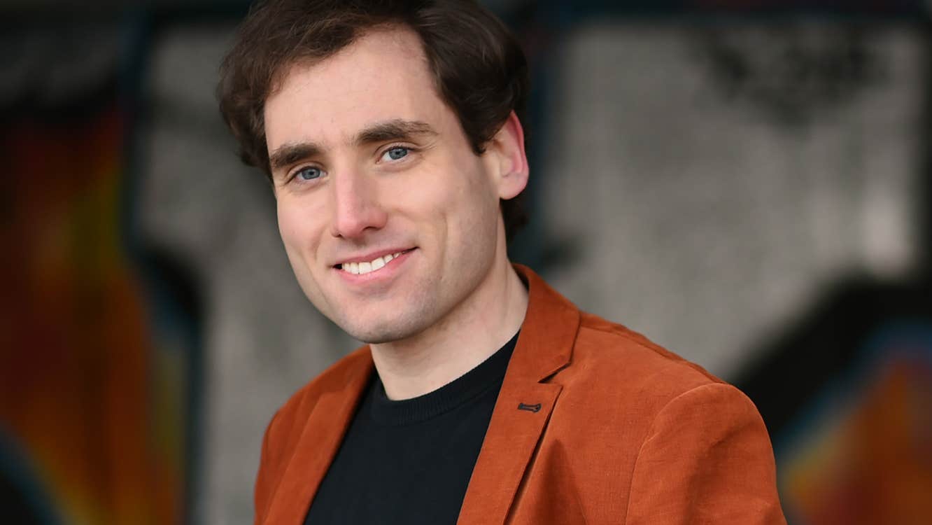 A man wearing a dark top and brown jacket is smiling at the camera.