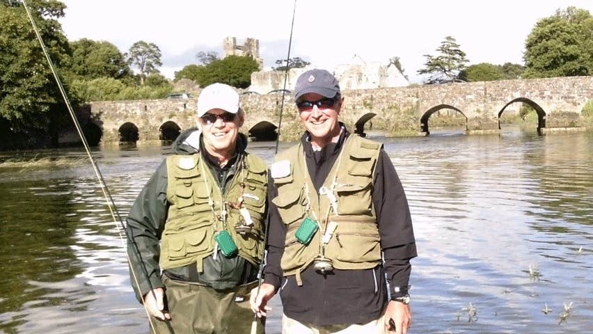 Two fishermen standing in the water holding fishing rods with a bridge in the background