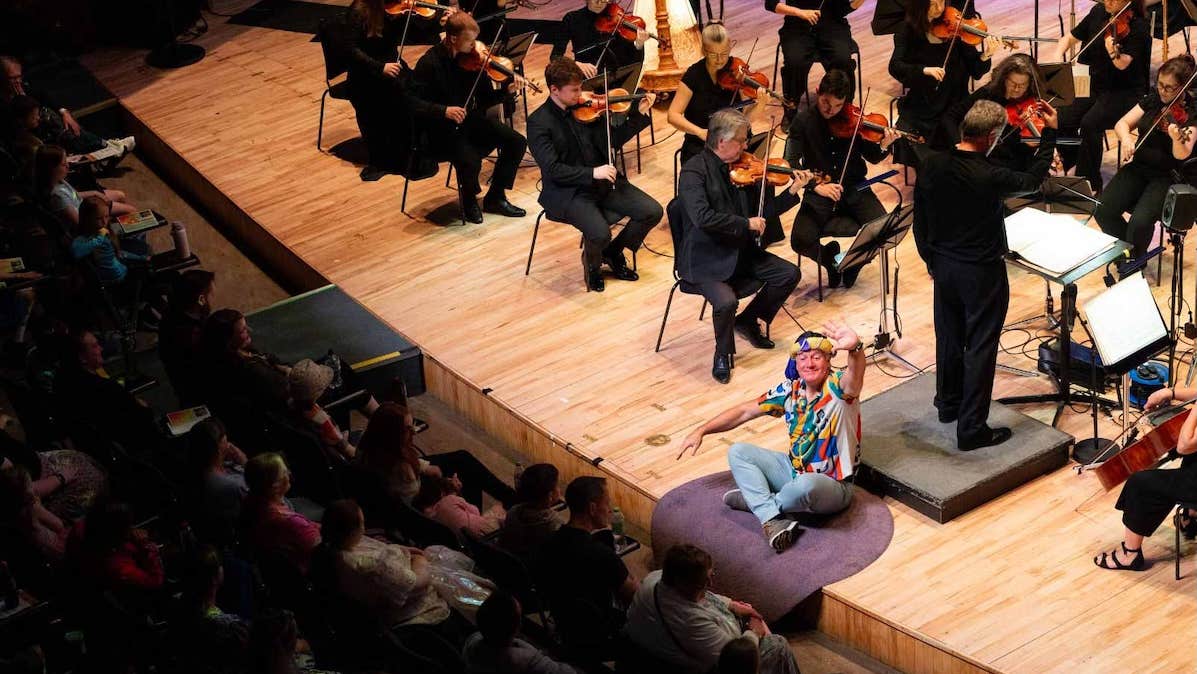 A smiling man in a colourful shirt is seated on the edge of a stage waving at the audience with an orchestra playing behind him.