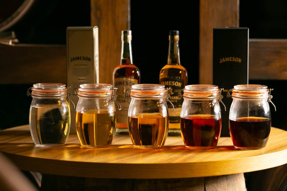 Five jars of different whiskies sit on top of a wooden table with Jameson bottles of whiskey standing behind.