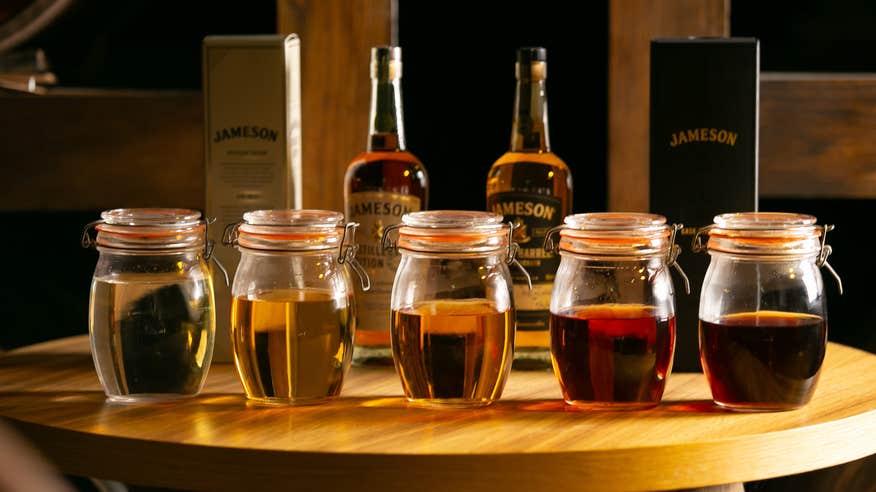 Five jars of different whiskies sit on top of a wooden table with Jameson bottles of whiskey standing behind.