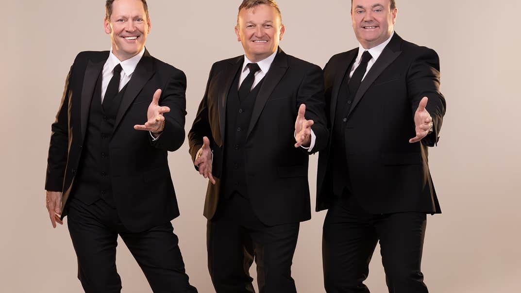 3 smiling men in black suits and ties each with their left hand outstretched, against plain beige background.