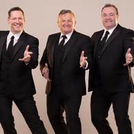 3 smiling men in black suits and ties each with their left hand outstretched, against plain beige background.