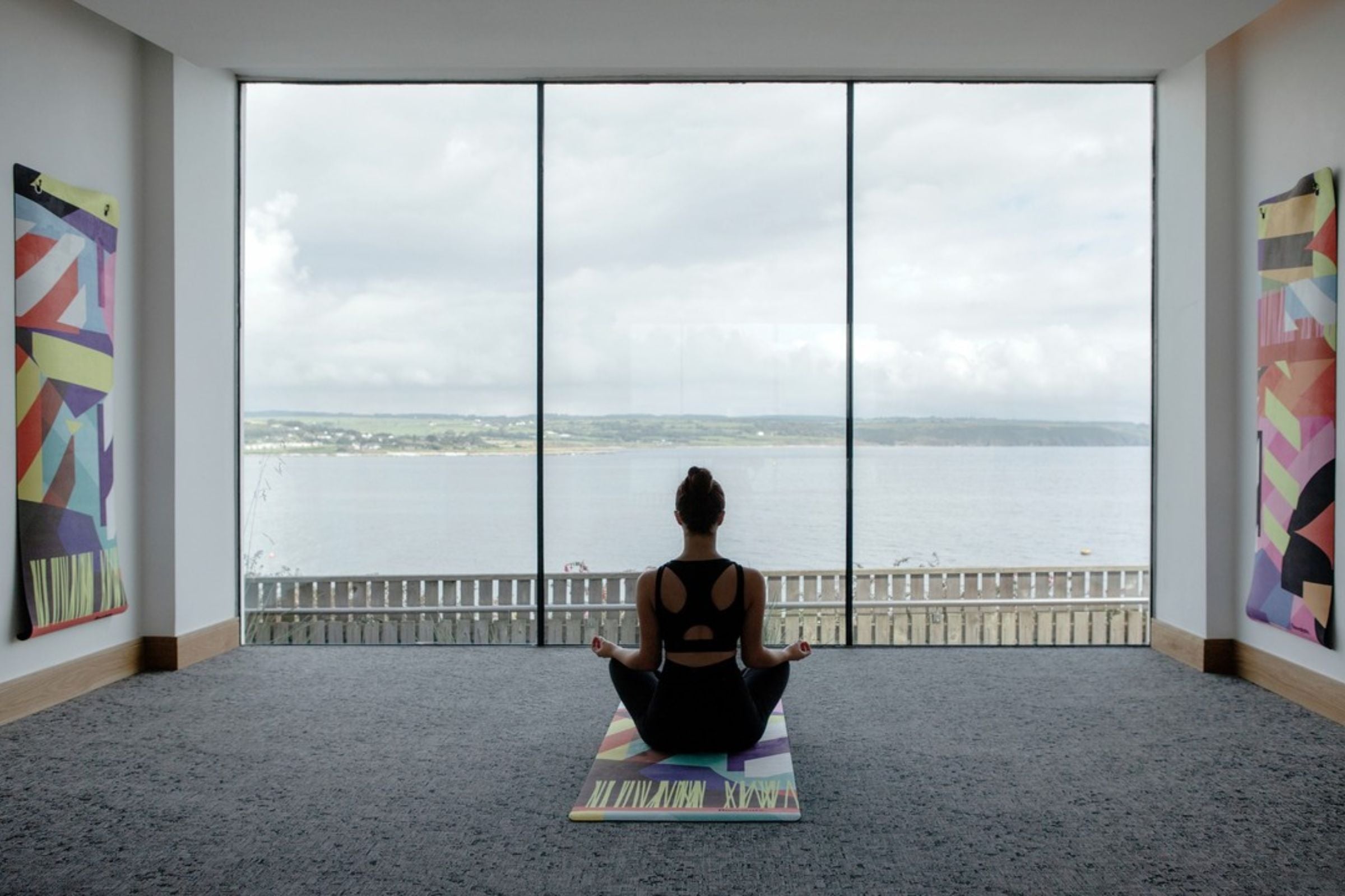 Woman sitting in a yoga pose infront of floor to ceiling windows with the ocean outside.
