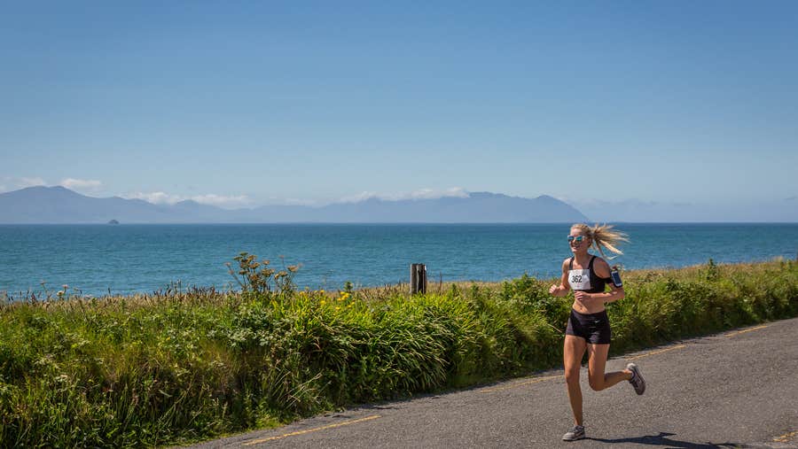 A sole figure running along a road with water and distant hills in the background and clear blue sky.