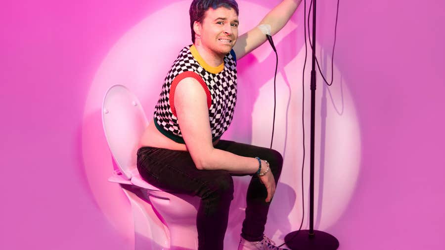 A grinning person seated, holding onto a mic on a stand with a bright pink background.