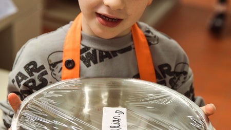 Child in an orange apron holding a silver bowl with a ball of dough in it