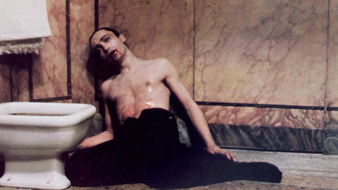 A shirtless man with waxy skin, with blood dripping from his mouth, is lying against a wall next to a toilet.
