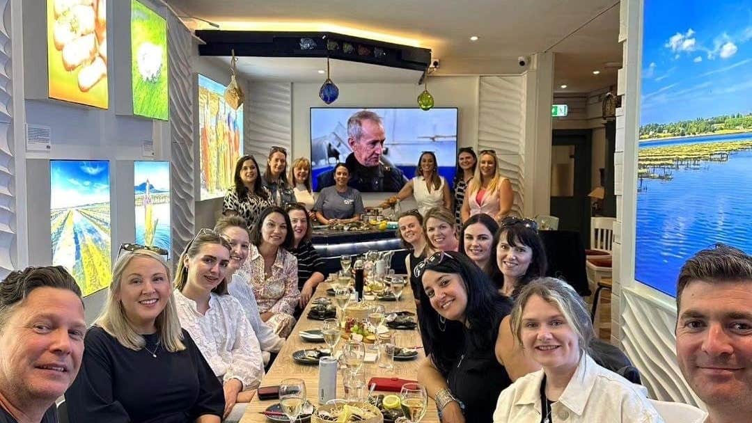 Group of people sitting at a long wooden table with food and wine glasses and bright screens on the walls