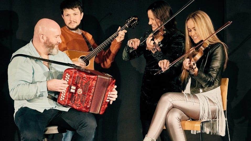 4 smiling musicians with their instruments, 2 seated with 2 standing behind, against plain, dark background.