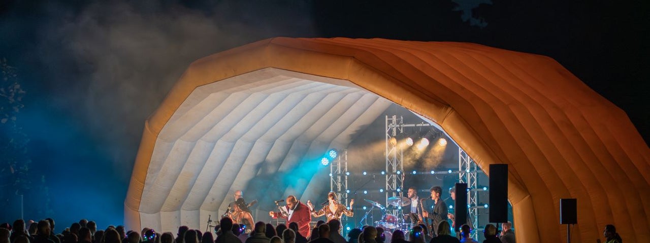 A blow up outdoor stage in orange with blue and white lights on performers at night with audience in darkness.