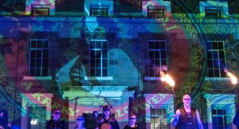 Samhain Chiarraí Street Spectacle, a line of people at night in costumes standing in front of a building lit up with logo from light show.