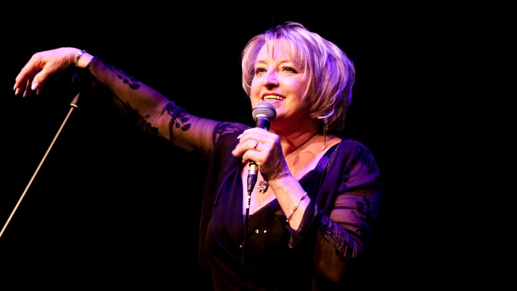 A woman on a dark stage singing into a mic she's holding with the other hand resting on a mic stand.