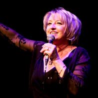 A woman on a dark stage singing into a mic she's holding with the other hand resting on a mic stand.