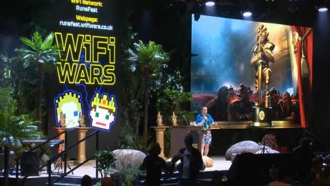 WiFi Wars: Interactive Comedy Game Show, view of a stage with a man talking, a large screen to one side.
