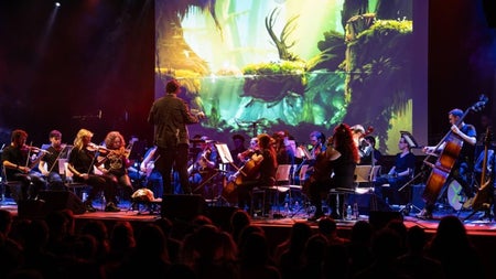Orchestra performing on stage with strings in the foreground and the conductor, large screen behind showing gameplay footage from the game. Audience silhouettes in foreground.