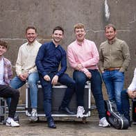 A group of men seated on large black equipment boxes with brown wall behind.