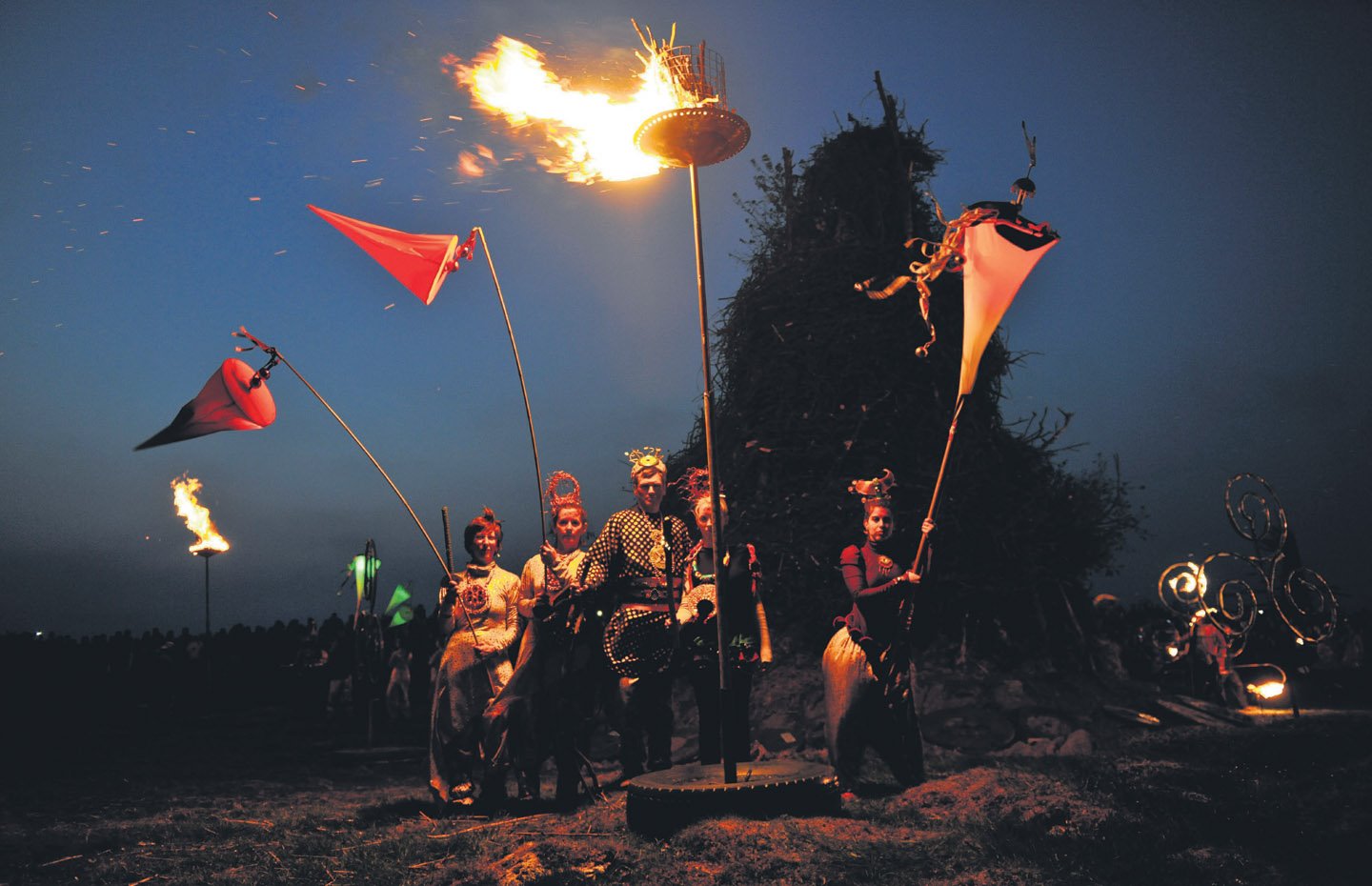 Bealtaine Fire Festival, Hill of Uisneach. At night, people in costume holding tall flags and flames are by a tall bush outdoors